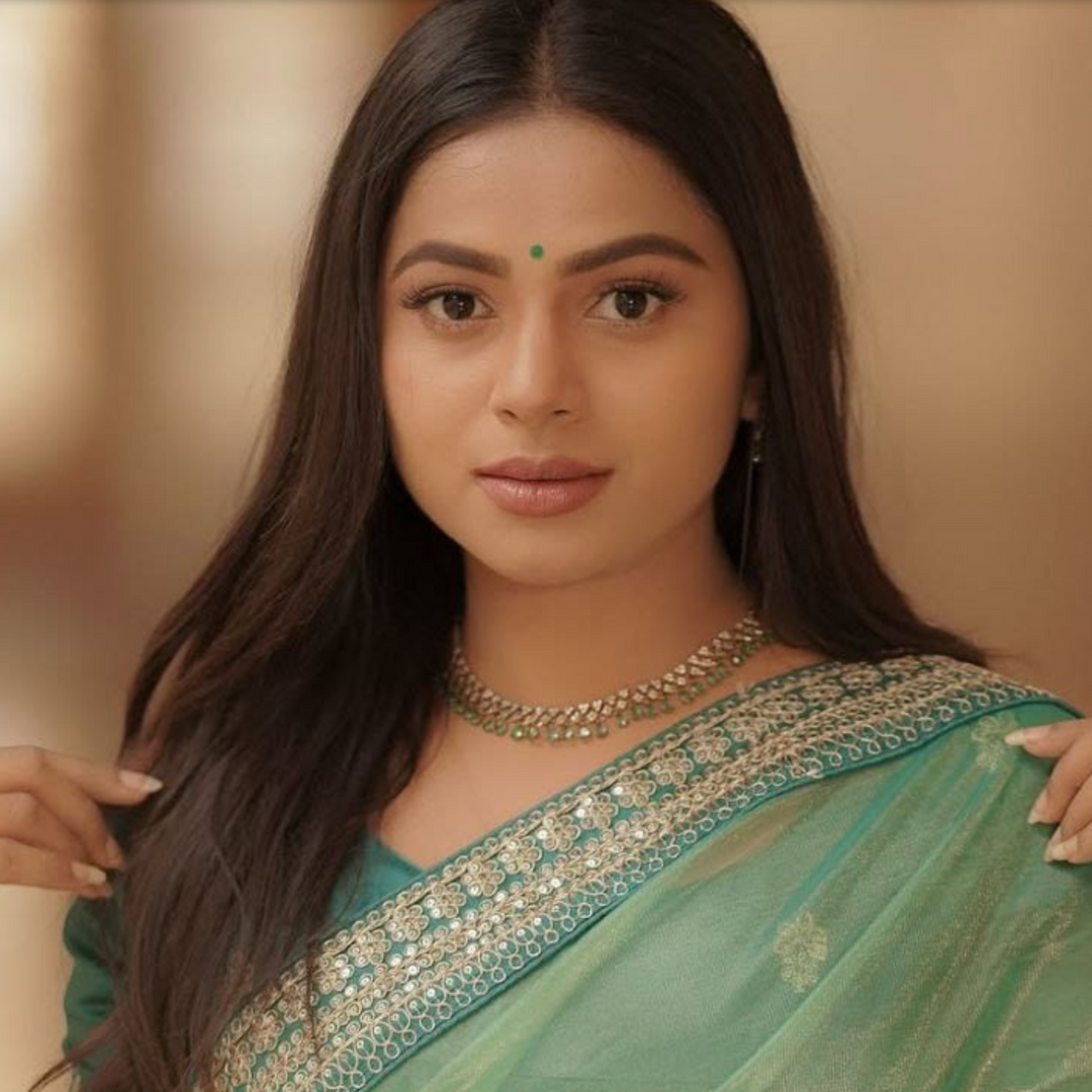 Woman wearing a green saree with intricate patterns, posing against a blurred background.