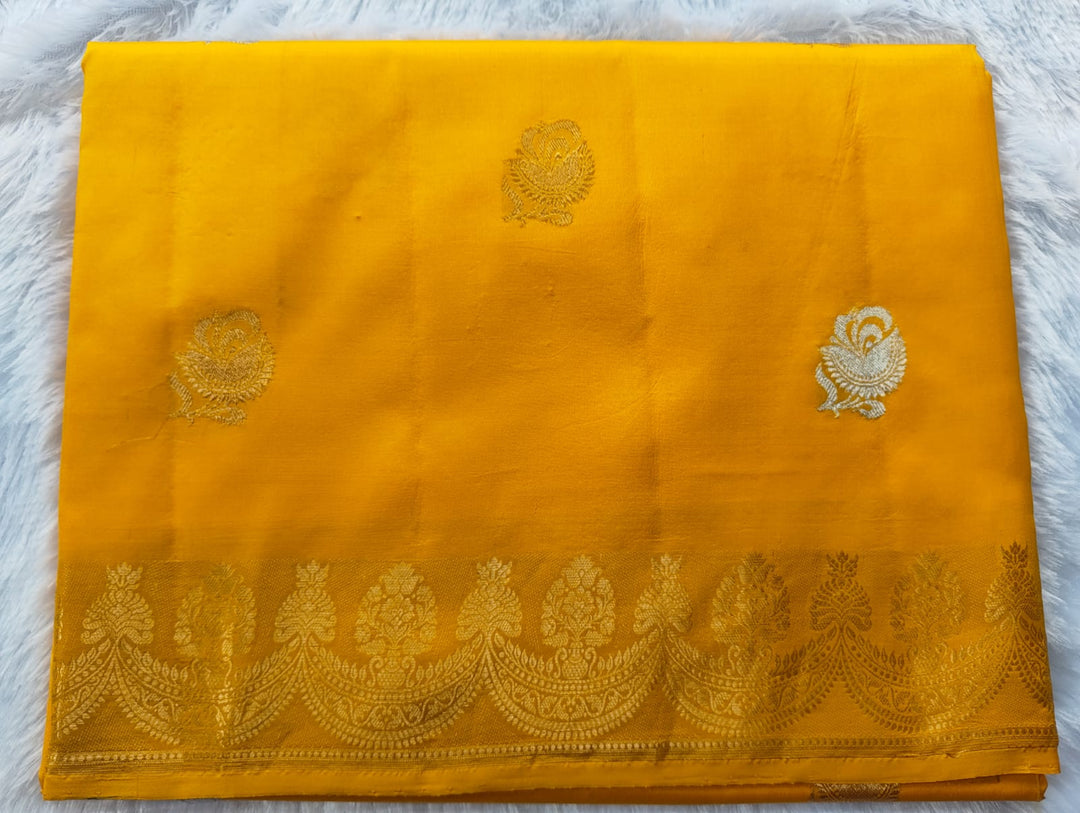 A folded yellow silk saree or fabric piece with a decorative gold zari border and small gold floral motifs, laid flat on a white textured background.